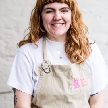 Alice Thatcher is a white woman with reddish brown hair. She has thick wavy hair and a fringe and is wearing a white top and a tan apron with a BCB logo on it.