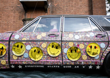 A car has been completely covered in a mosaic of pink tiles with yellow smiley face accents as part of Mad in Stoke by Carrie Reichardt. Displayed at the 2023 British Ceramics Biennial.