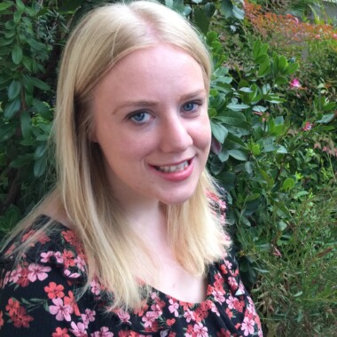 Photo shows a smiling white woman in her early thirties with shoulder length blonde hair. She is wearing a black and orange floral top and is standing in front of a bright green bush.