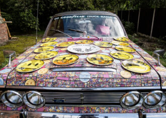 A car has been completely covered in a mosaic of pink tiles with yellow smiley face accents as part of Mad in Stoke by Carrie Reichardt. Displayed at the 2023 British Ceramics Biennial.